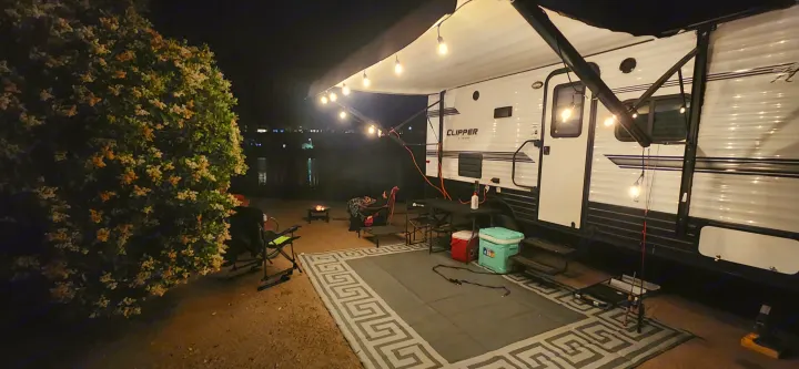Lakeside rv rental setup at night: glowing string lights, comfy chairs, and a warm fire pit create a perfect camping ambiance. Coachmen Clipper 2018