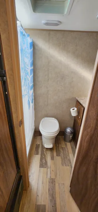 Inside an RV rental bathroom, featuring a toilet, shower with blue leaf curtain, and wood-look floor. Compact & functional.. Coachmen Clipper 2018
