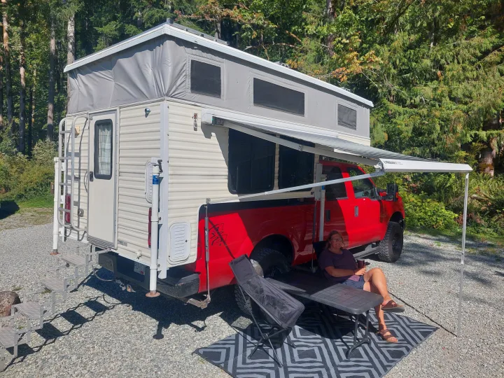 Relaxing RV rental setup: a red truck camper with extended awning provides shade for a person enjoying the outdoors.. Ford F350 2015