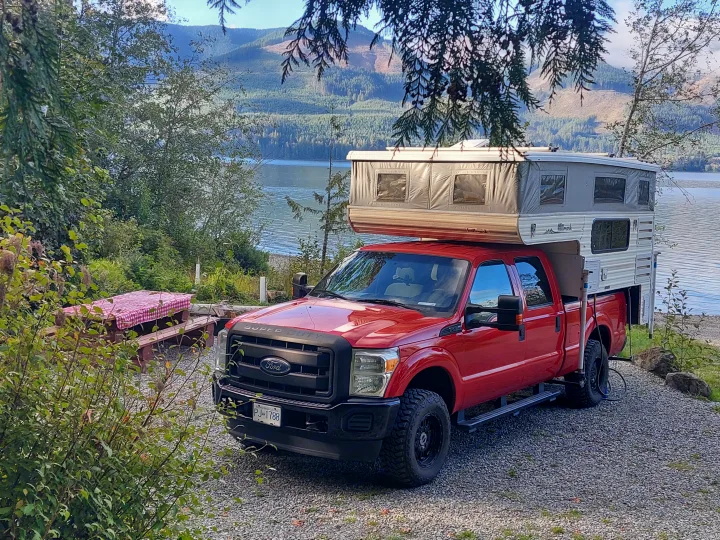 Red Ford truck with pop-up truck camper RV rental parked by a scenic lake, ideal for outdoor camping adventures.. Ford F350 2015