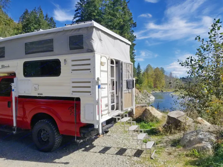 Scenic RV rental setup by a serene river, showcasing a pop-up truck camper and outdoor steps under a clear sky.. Ford F350 2015