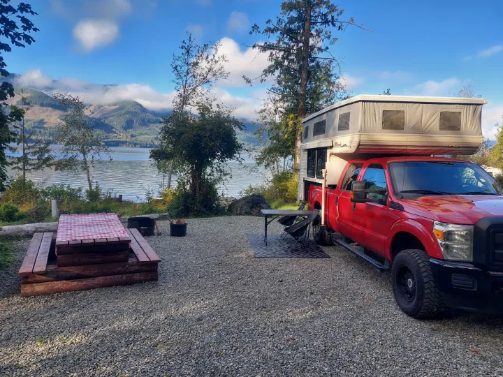 Perfect rv rental getaway! Red truck camper at a lakeside campsite with a picnic table & cloudy mountains.. Ford F350 2015
