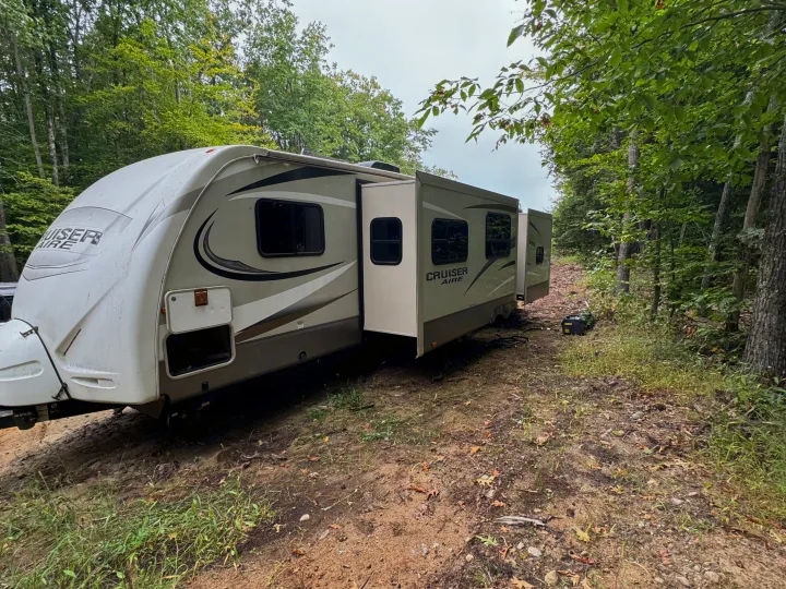 An RV rental, a travel trailer, is parked on a dirt path in a wooded setting, complete with a portable generator.. Crossroads RV Cruiser Aire 2016