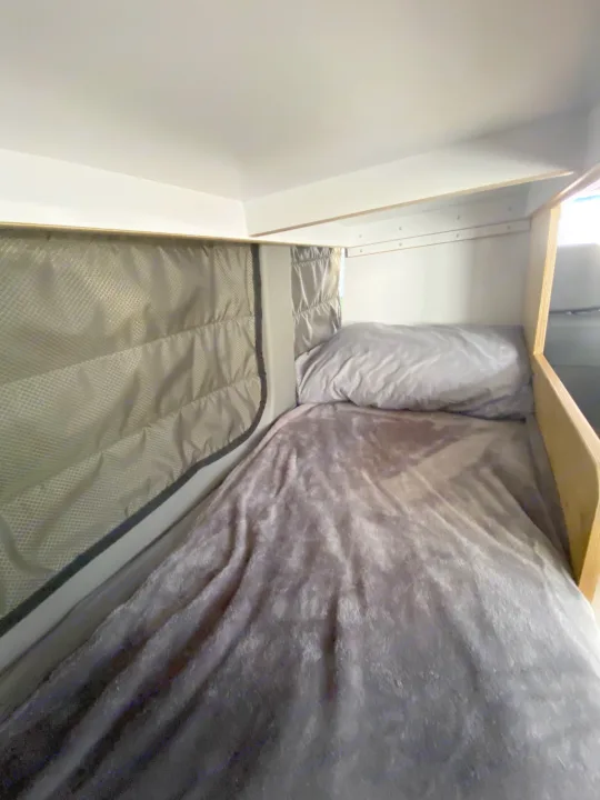Inside an RV rental: a compact and comfortable sleeping area with a gray bed, pillow, and padded window cover.. Mercedes-Benz Sprinter 2014
