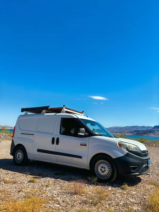 White adventure van with roof rack, awning, by a lake & mountains under clear blue sky. Perfect RV rental.. Dodge Promaster City 2016
