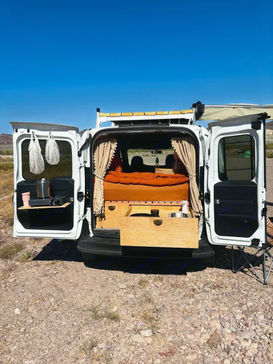 A white **rv rental** camper van with open rear doors reveals a custom interior: bed, kitchen, and wooden storage. Perfect for outdoor adventures.. Dodge Promaster City 2016