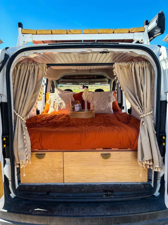 View inside a stylish RV rental van conversion: comfy orange bed, wood storage, and decorative curtains.. Dodge Promaster City 2016