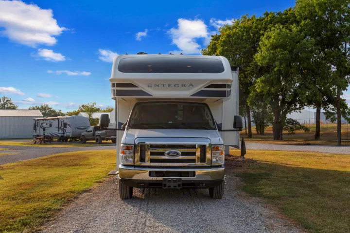 Front view of a white Ford Class C RV rental, Entegra brand, parked on a gravel lot. Trees and other RVs visible.. Entegra Coach Odyssey 2020