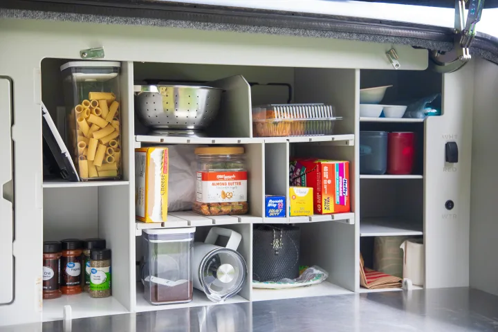 Organized RV rental kitchen pantry with shelves holding pasta, snacks, spices, cooking tools, and supplies for camping.. Escapod Topo 2 Voyager 2024