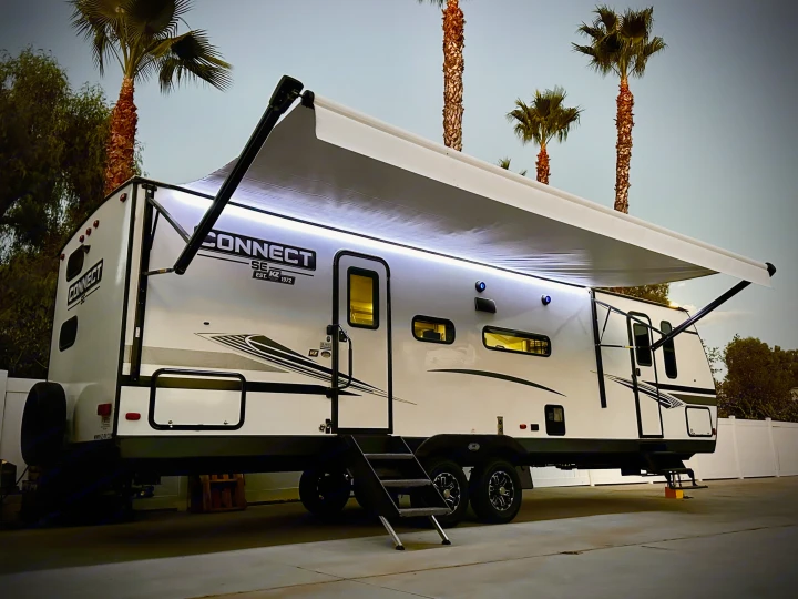 Modern white RV rental travel trailer with bright awning lights, parked under palm trees at dusk. Ready for adventure.. Connect SE 2022