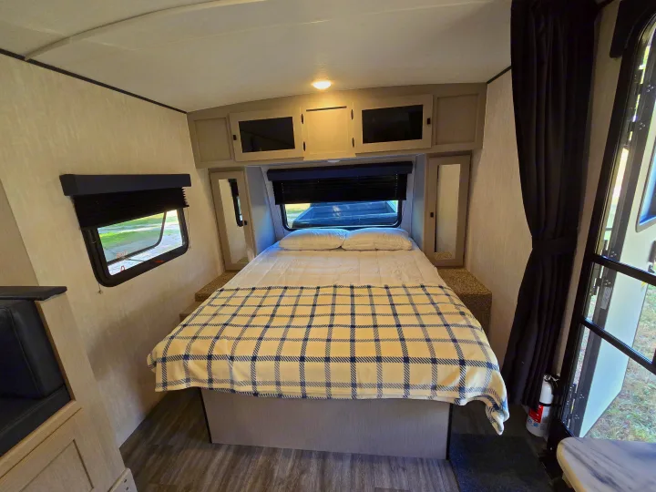 This compact RV rental bedroom features a double bed with a plaid blanket, overhead cabinets, and windows with blinds.. Coachmen Apex Nano 2024