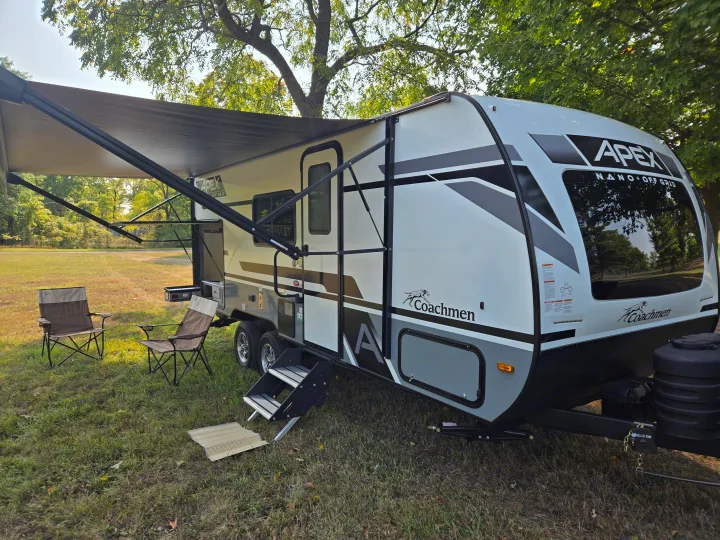 This white and grey travel trailer, an RV rental, is set up with an extended awning and camping chairs in a grassy, shaded area.. Coachmen Apex Nano 2024