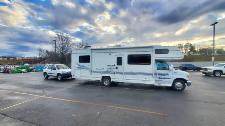 This white RV, seen towing an SUV in a cloudy parking lot, is typical of an rv rental for road trips.. Coachmen Santara 1998