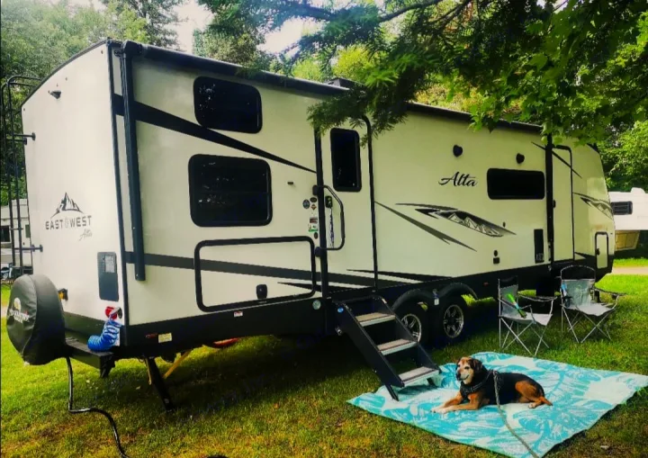 Relaxing RV rental setup: a white travel trailer under trees, with a happy dog on a mat, perfect for camping.. East to West Alta 2022