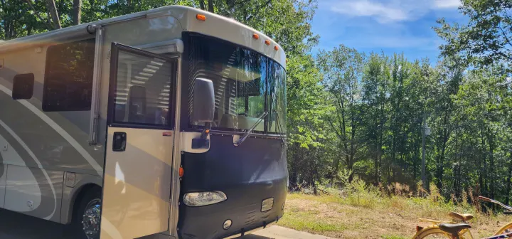 A spacious RV rental, light-colored with an open door, is parked in a sunny, wooded area. Ideal for your next outdoor adventure.. Winnebago Journey 2006