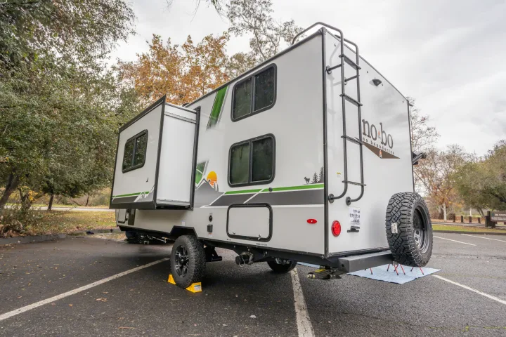 A white no.bo RV rental travel trailer with slide-out and spare tire, parked on asphalt with chocks.. No Boundaries 19 2023