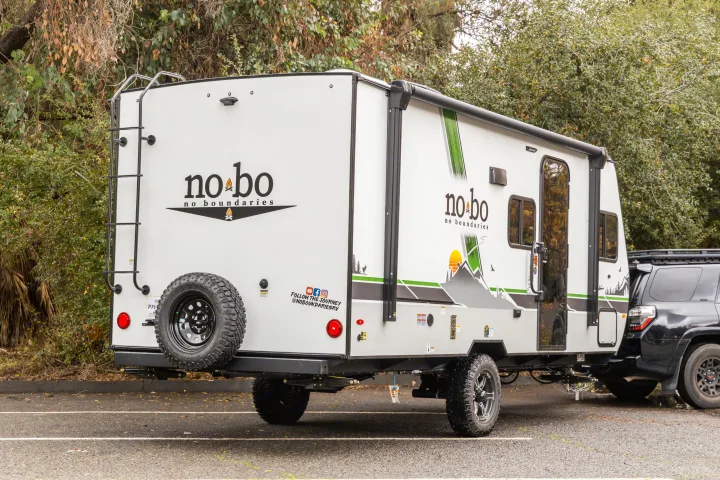 A white 'No Boundaries' travel trailer for rv rental, ready for camping & outdoor adventures. Parked with an SUV.. No Boundaries 19 2023
