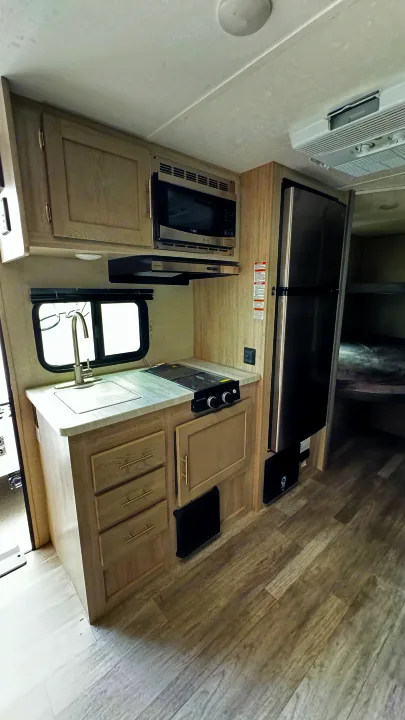 RV rental interior featuring a compact kitchen with microwave, sink, stove, fridge, and bunk beds in the background.. Coachmen Catalina 2023