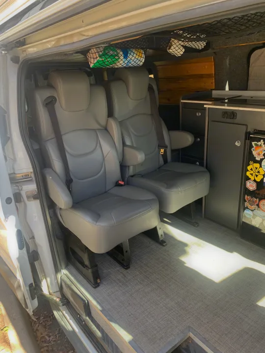 Inside an RV rental: two grey passenger seats with seatbelts, a kitchenette, and overhead storage nets.. Ford Transit Custom 2019