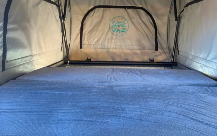 Interior of a pop-top RV rental, featuring a comfortable grey bed and "Colorado Camper Van" logo visible on the back fabric.. Ford Transit Custom 2019