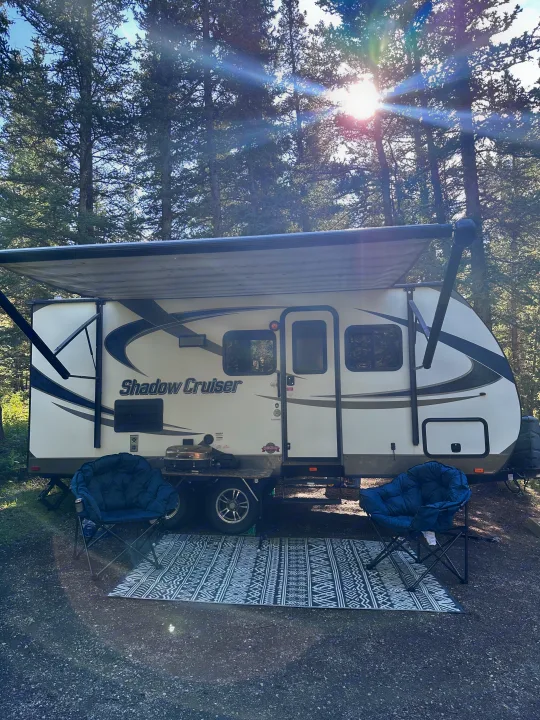 Peaceful RV rental: travel trailer with deployed awning, chairs, and grill. Sunny forest campsite setup.. Cruiser RV Shadow Cruiser 2016
