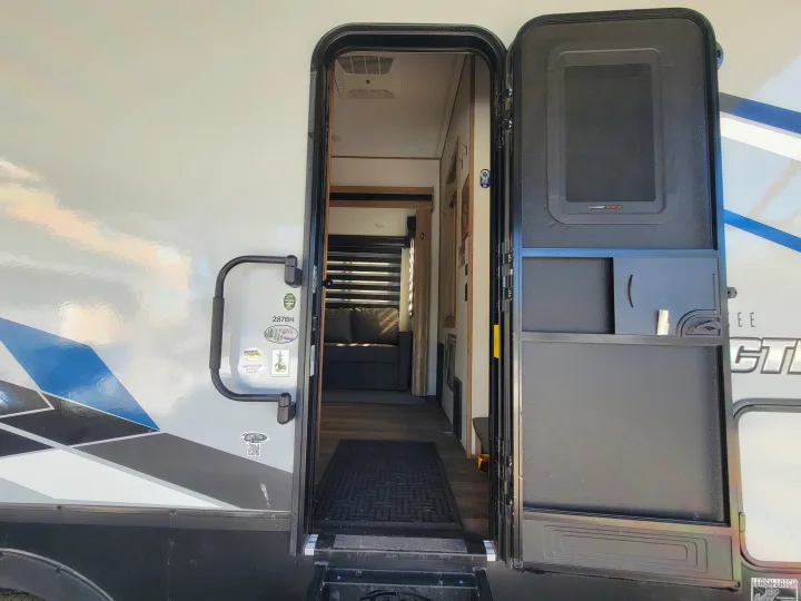 An open RV rental door reveals inside steps, a floor mat, and a grey couch. The exterior has a large grab handle.. Forest River Cherokee Arctic Wolf 2023