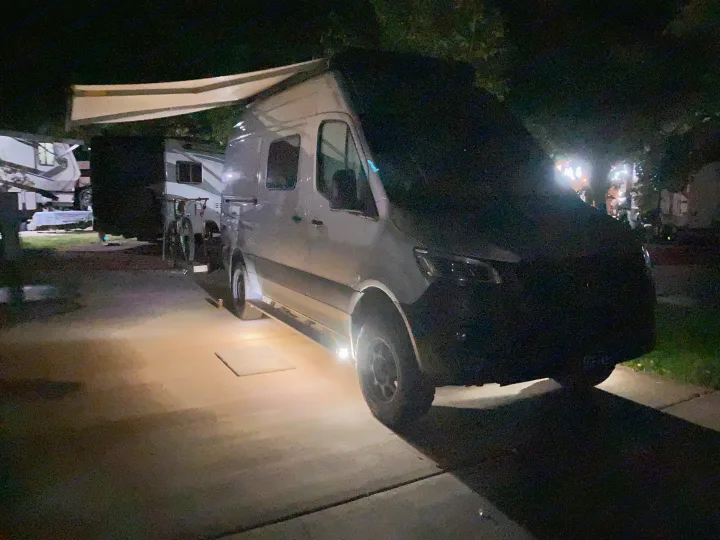 A white camper van, a common RV rental, is parked at a campsite at night, with an awning extended and under-vehicle lights on.. Winnebago Revel 2020