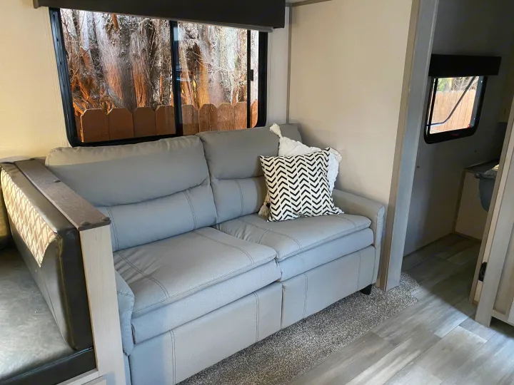 Inside an RV rental: a comfortable gray couch with pillows and large windows showing trees and a fence.. Forest River Tracer 2021