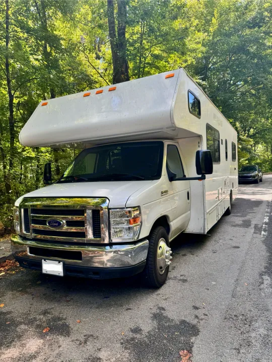 An RV rental, a white Ford E-450 motorhome, is parked on a shaded, tree-lined road, ready for travel.. Thor Majestic 2019