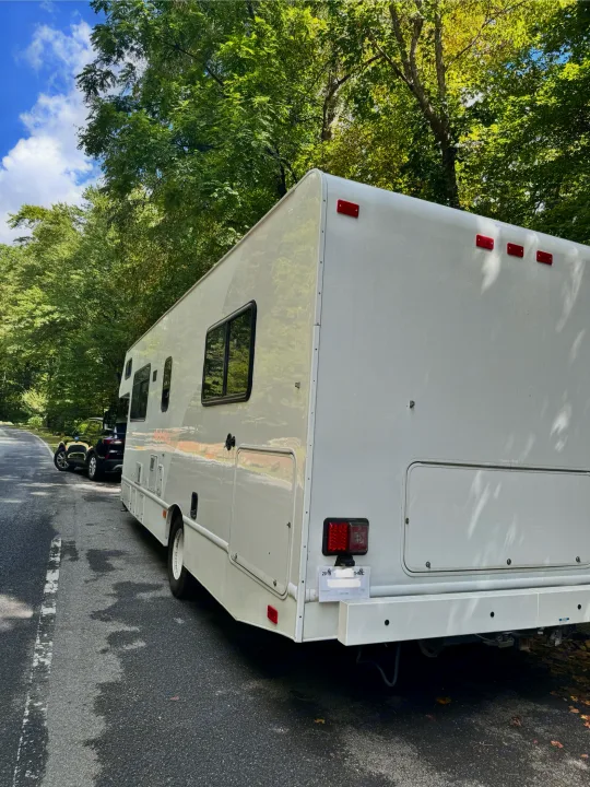 Large white RV rental parked curbside on a scenic, tree-lined road. A small car is visible further ahead.. Thor Majestic 2019