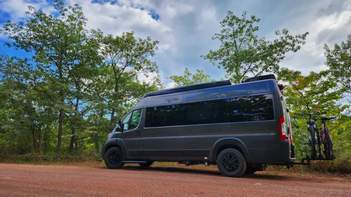 Dark grey RV rental with bikes on back, parked on dirt road in green forest. Perfect for nature adventures.. Airstream Rangeline 2024