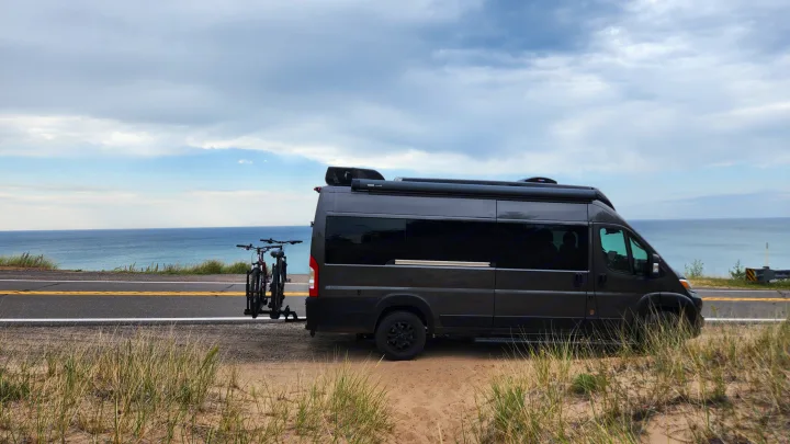 A dark grey RV rental, a camper van, parked on a sandy roadside next to a large blue body of water, with bikes on back.. Airstream Rangeline 2024