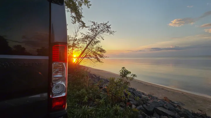 RV rental's serene sunset: A camper van overlooks a calm beach with trees & a vibrant horizon.. Airstream Rangeline 2024