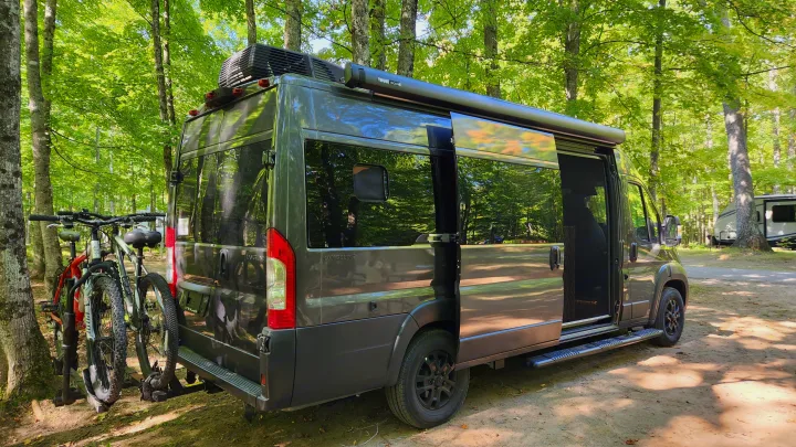 RV rental: Dark grey camper van with bikes on the rack & open side door, parked at a lush green campsite.. Airstream Rangeline 2024
