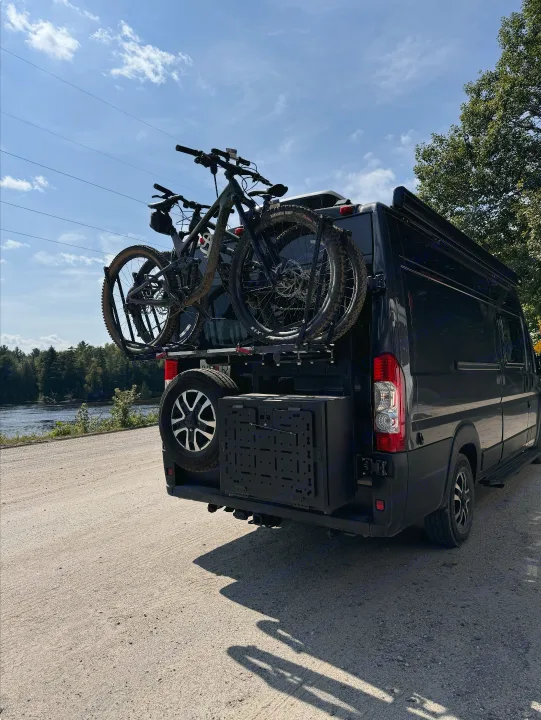 Black camper van on dirt road, carrying two mountain bikes & spare tire. Ideal for an rv rental adventure!. Noovo Pop 2024