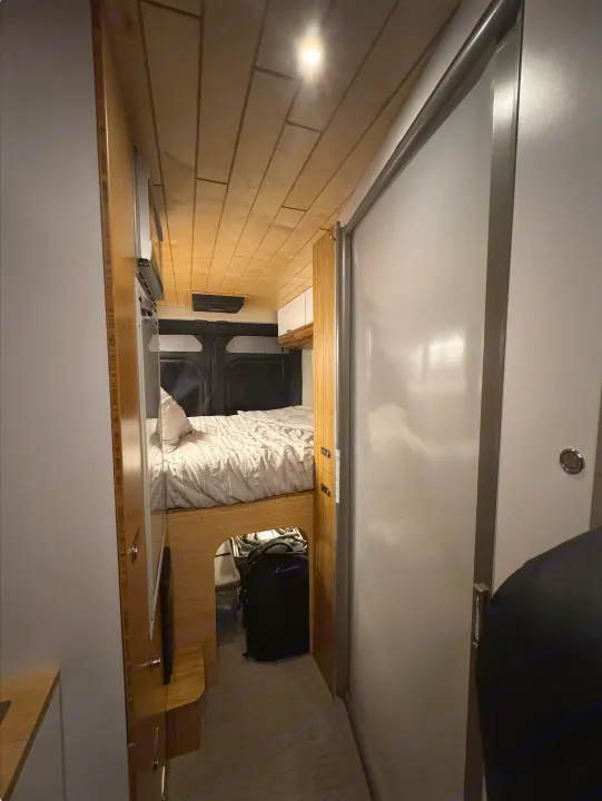 Inside an RV rental: a modern, narrow interior with a cozy bed, under-bed storage, wooden cabinetry, and a light sliding door.. Noovo Pop 2024