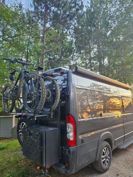RV rental ready: Grey camper van with a rear bike rack holding two mountain bikes, parked by trees.. Noovo Pop 2024