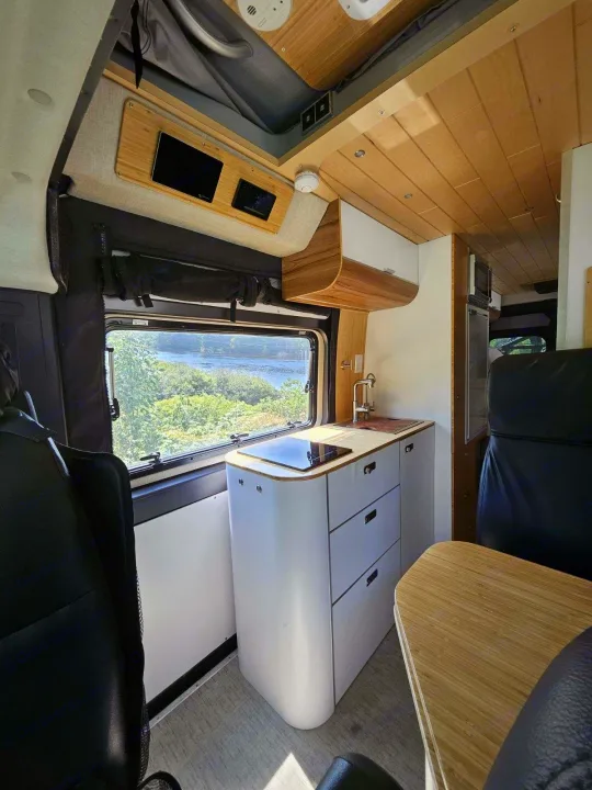 Bright rv rental interior: compact kitchen, large window with scenic river view, and wood accents for travel.. Noovo Pop 2024