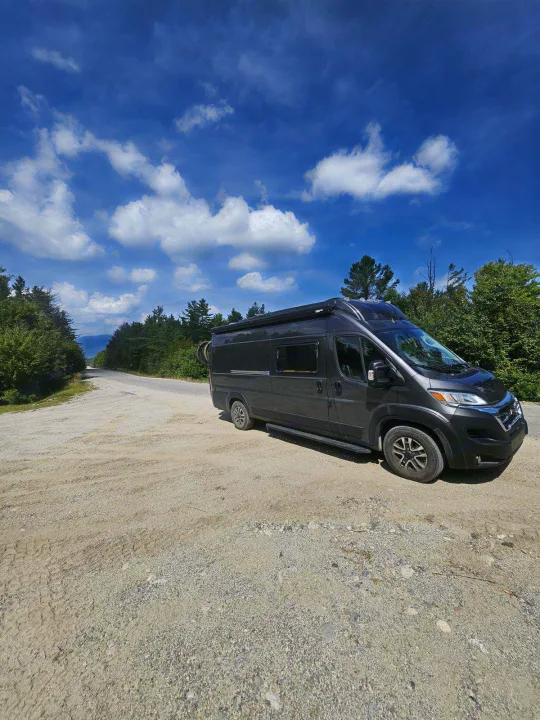Explore with an RV rental! Dark grey camper van parked on a scenic gravel road amidst lush trees and blue sky.. Noovo Pop 2024