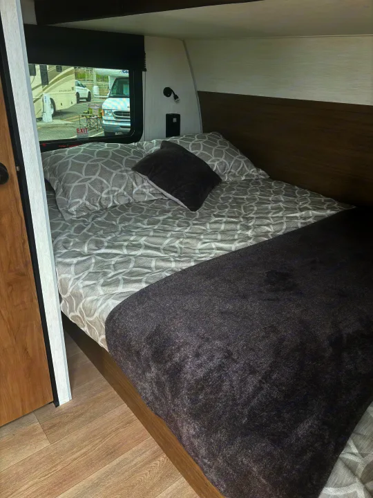 Cozy RV rental bedroom with patterned bed, dark blanket, window view of other vehicles, plus a light and outlet.. Forest River Wolf Pup 2025