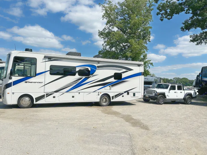 A large white WindSport motorhome, an ideal rv rental, is parked on a gravel lot with a white Jeep Gladiator.. Thor Windsport 2023
