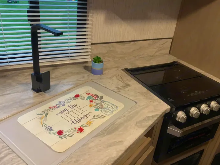 This RV rental kitchen features a modern black faucet, stove, and a counter with a cutting board saying 'enjoy the little things'.. Heartland RVs Sundance Ultra Lite 2021