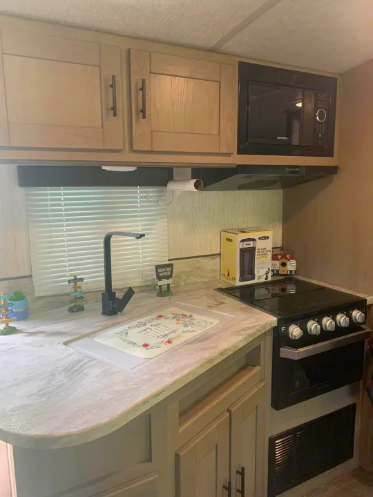 This RV rental's modern kitchen features light cabinets, black appliances (microwave, oven), and a sleek faucet on light counters.. Heartland RVs Sundance Ultra Lite 2021