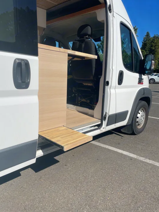 This RV rental shows a white camper van with an open side door, revealing its custom interior and a pull-out wooden table.. Vanberry Promaster 2018