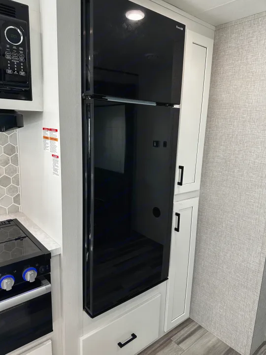 This compact RV rental kitchen features modern black appliances, including an Everchill refrigerator, microwave, and stovetop, alongside sleek white cabinetry.. Jayco Jay Flight 2024