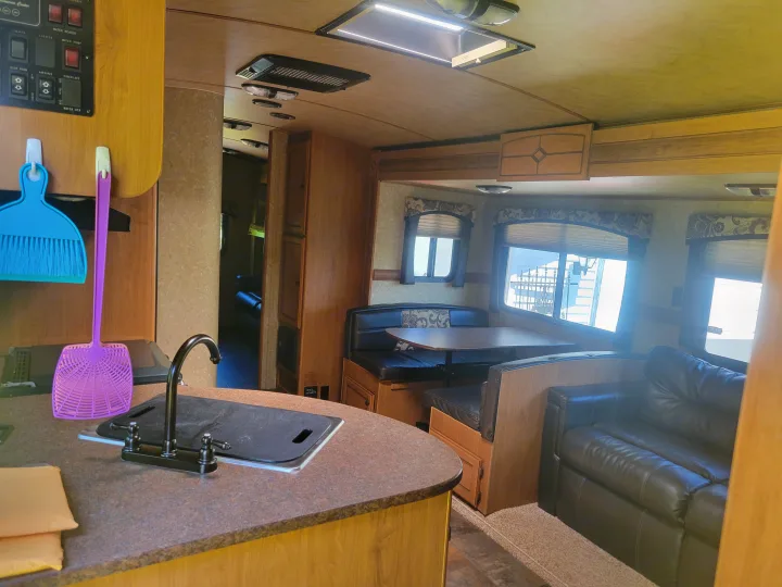 RV rental interior: kitchen counter with sink, dinette with dark booths, and a comfy sofa. Controls and bright windows are visible.. Crossroads RV Sunset Trail Super Lite 2015