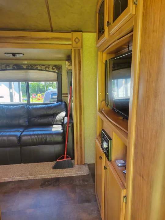 This RV rental's living space features a black leather sofa, entertainment center with TV, and window view. A broom is by the couch.. Crossroads RV Sunset Trail Super Lite 2015