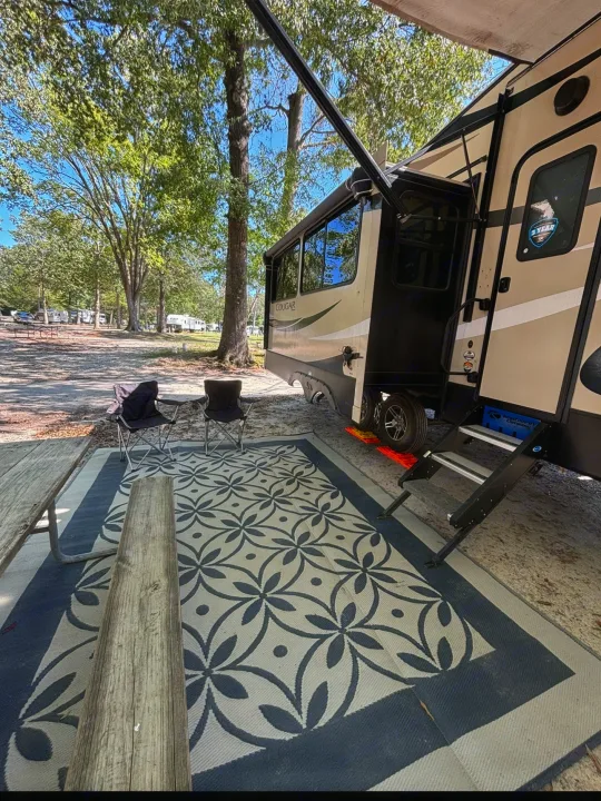 An RV rental set up for comfort with an extended awning, outdoor rug, camping chairs, and picnic table at a shaded campground.. Keystone RV Cougar 2019