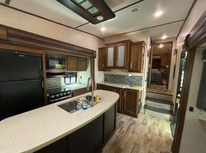 Spacious RV rental kitchen with fridge, stove, microwave, and island sink. Steps lead to a cozy bedroom.. Keystone RV Cougar 2019