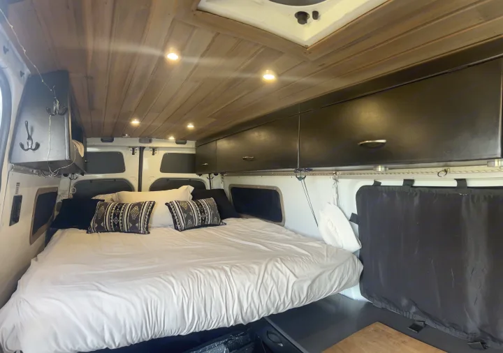 Cozy RV rental interior with a comfortable bed, wooden ceiling lights, and overhead storage, perfect for mobile travel.. Freightliner Sprinter 2016